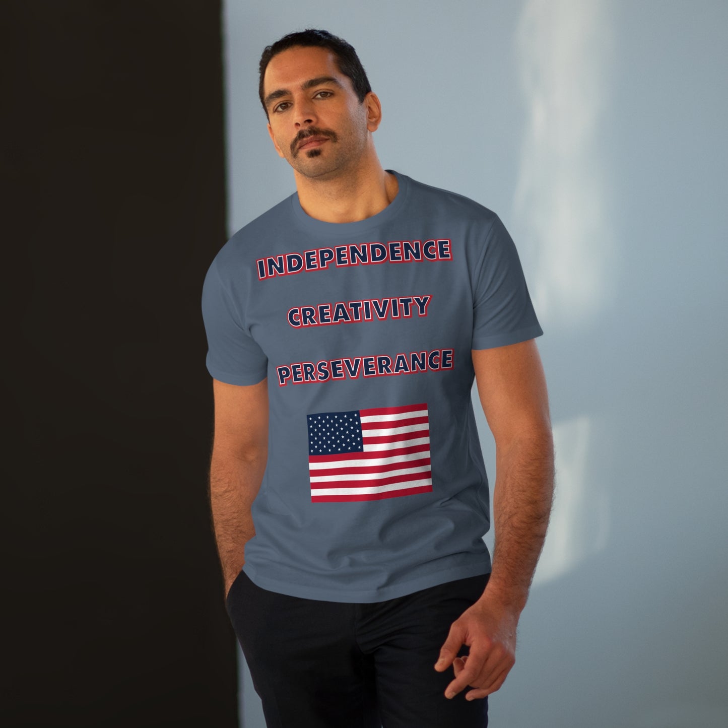 A man with a mustache wearing a mens grey t-shirt with the words independence, creativity, and perseverance written in bold, red, white, and blue letters, with an American flag located below.