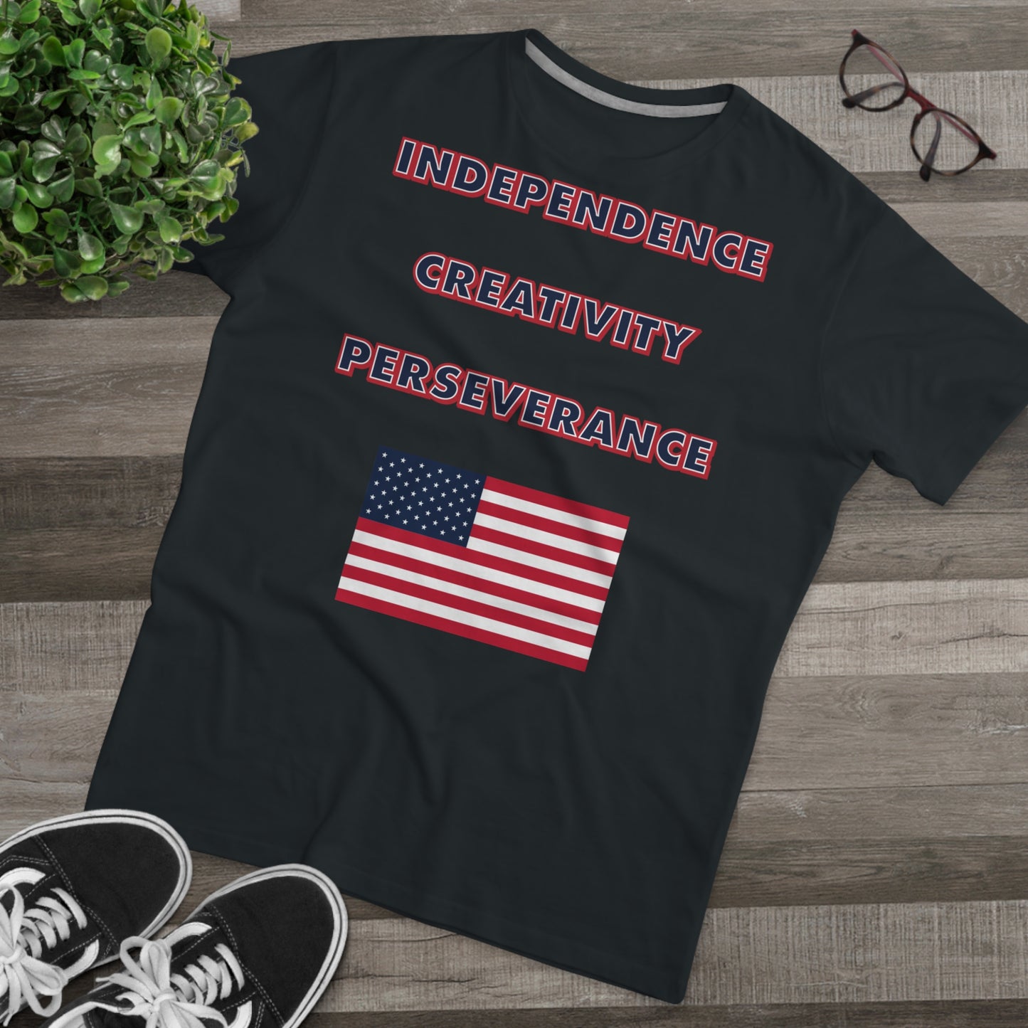 A mens black t-shirt with the words independence, creativity, and perseverance written in bold, red, white, and blue letters, with an American flag located below. The shirt is laid on a hard wood floor next to a pair of brown glasses, a green house plant, and a pair of black canvas sneakers.