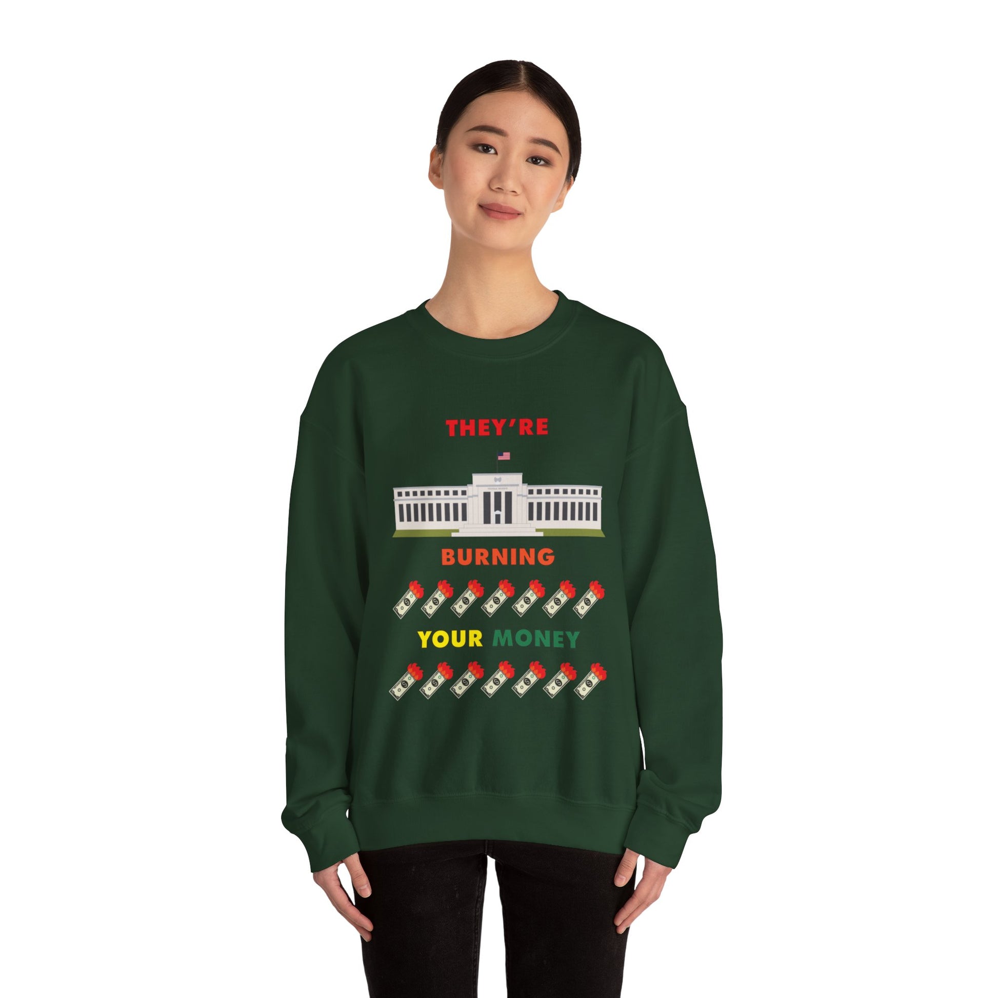 A woman wearing a green crewneck sweatshirt with the words "They're Burning Your Money" in large bold letters. The design has a digital drawing of the United States Federal reserve building in Washington D.C. and it features drawings of burning dollar bills.