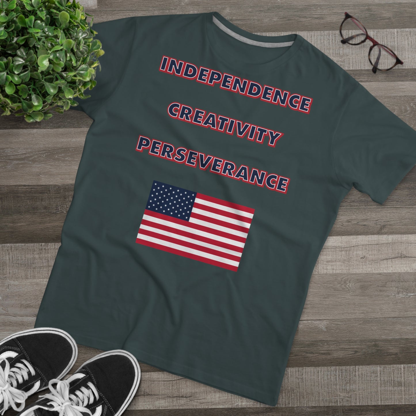 A charcoal grey t-shirt with the words independence, creativity, and perseverance written in bold, red, white, and blue letters, with an American flag located below. The t-shirt is laid on top of a hard wood floor next to a pair of glasses and a pair of black shoes.